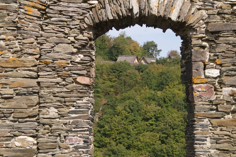 Burgmauern, Fenster, Mauerdurchbruch, und durch das ehemalige Fenster sieht man die Hütten der rekonstruierten Keltensiedlung.