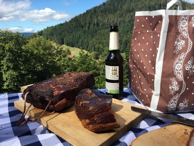Tisch mit Schinken und Bier, dahinter die grüne Schwarzwald-Szenerie mit Wald und blauem Himmel.