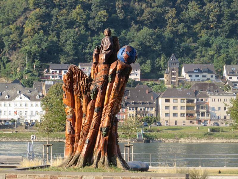 Eine bunte Holzskuptur am Fluss, am gegenüberliegenden Ufer sieht man die Häuser einer Ortschaft