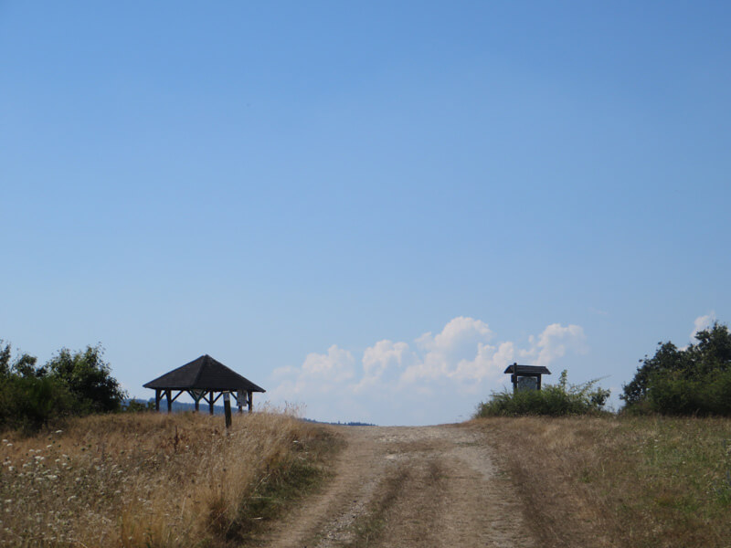 Ein weiße Quellwolke am sommerlichen HImmel. Fast nur als Schattenriss erkennbar: eine Schutzhütte links vom Weg, auf der rechten Seite eine Infotafel. 