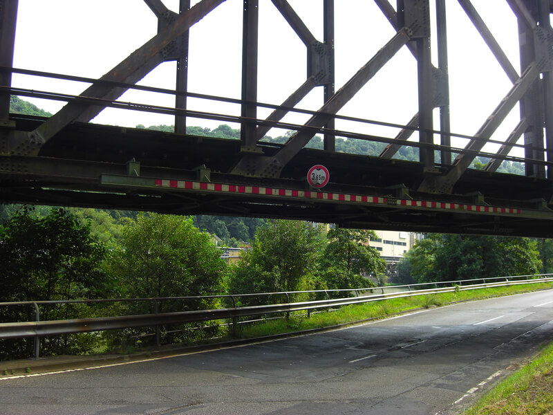 unten eine Straße begrenzt von grünem Gebüsch, darüber die Eisenkonstruktion einer alten Eisenbahnbrücke, die Hohenrheinbrücke bei Niederlahnstein
