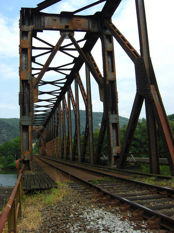Das rostige Eisengerüst eines alten Bahnviadukts rahmt eine eingleisige Bahnstrecke ein. 