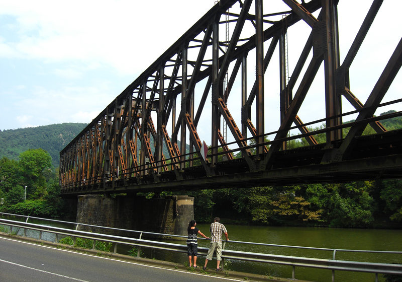 Ein rostiges Stahlfachwerk über einen Fluss, davor zwei Menschen, die sich das Wasser des Flusses anschauen